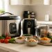 Cuisine lumineuse avec un multicuiseur, un air fryer, un panier vapeur en inox, des légumes frais et un repas maison sain sur le plan de travail.